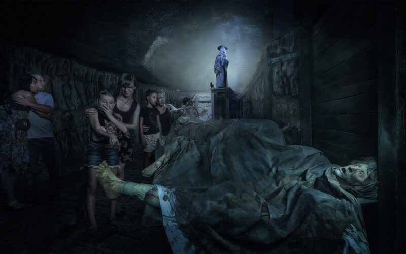 Eine Gruppe von Menschen geht durch einen dunklen, unheimlichen Raum mit bedeckten Körpern auf dem Boden, während eine gewandete Gestalt auf einem Podest am anderen Ende des Raumes steht und leuchtet.