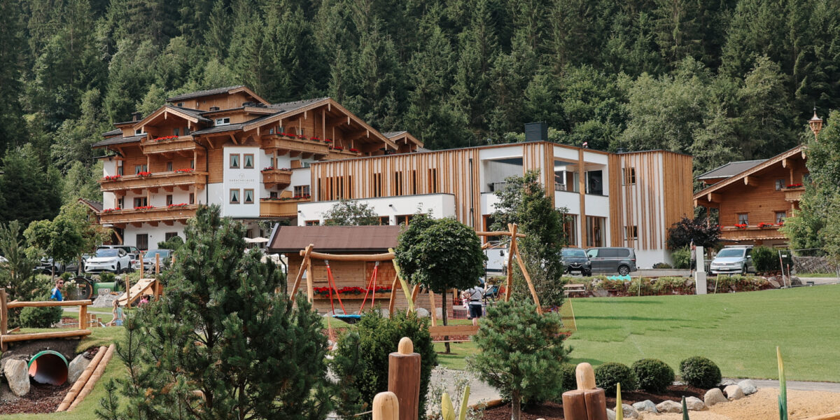 Ein modernes Hotel und Gebäude im Chalet-Stil, umgeben von Bäumen, mit einem Spielplatz und Landschaftsgärten im Vordergrund.