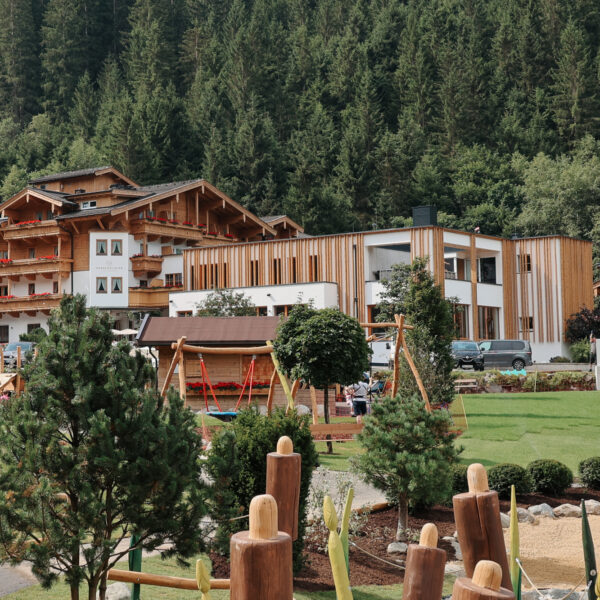 Ein modernes Hotel und Gebäude im Chalet-Stil, umgeben von Bäumen, mit einem Spielplatz und Landschaftsgärten im Vordergrund.