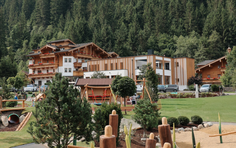 Ein modernes Hotel und Gebäude im Chalet-Stil, umgeben von Bäumen, mit einem Spielplatz und Landschaftsgärten im Vordergrund.
