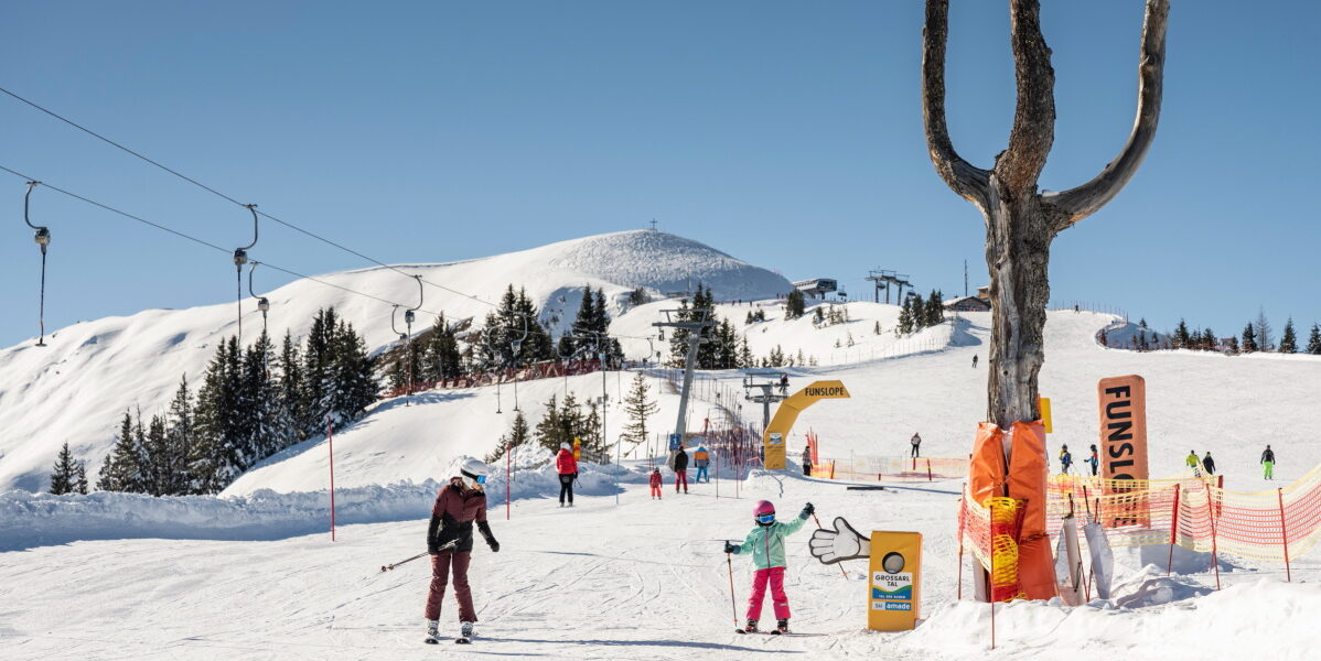 Skifahrer befinden sich auf einem verschneiten Berghang in der Nähe eines Skilifts und einer großen Holzskulptur in Form eines Dreizacks. Strahlend sonniges Wetter mit klarem blauen Himmel.