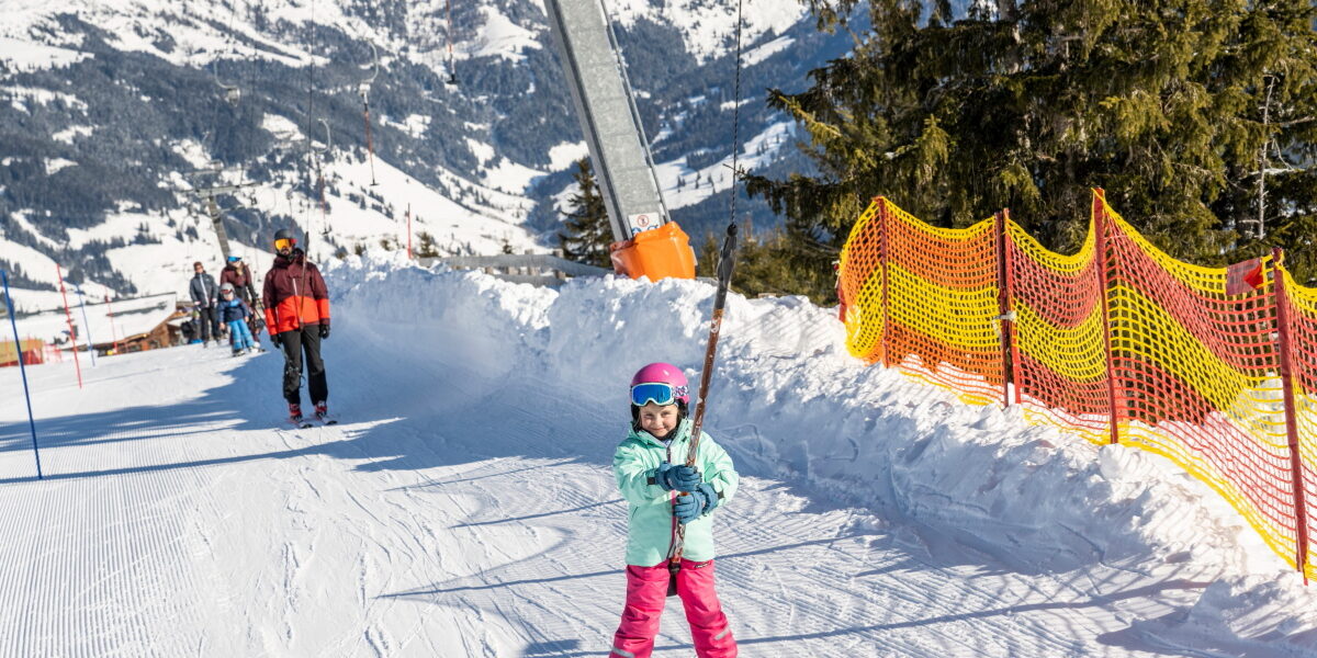 Ein Kind mit Helm und Skiausrüstung benutzt einen Skilift auf einer verschneiten Piste, mit Bergen und anderen Skifahrern im Hintergrund.