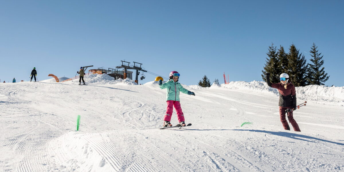 Zwei Kinder fahren auf einer verschneiten Piste mit Skistöcken Ski, tragen Helme und Winterausrüstung; im Hintergrund sind andere Skifahrer und ein Skilift bei klarem blauen Himmel zu sehen.