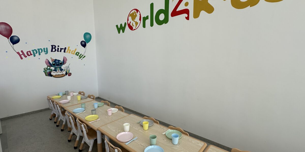 Ein Raum für Kinderpartys mit zwei langen Tischen, die mit pastellfarbenen Tellern und Tassen gedeckt sind. An der Wand hängen "world4kids"- und "Happy Birthday"-Dekorationen.
