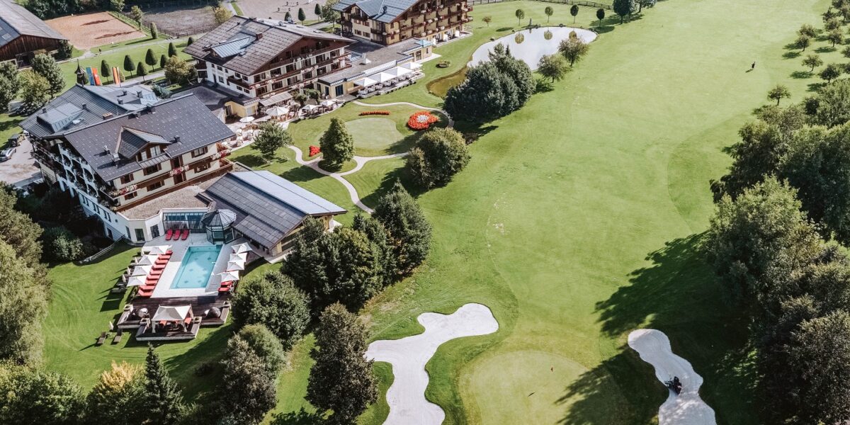 Luftaufnahme eines Resorts mit einem Pool, umliegenden Gärten, kleinen Teichen und einem angrenzenden Golfplatz mit Sandbunkern und grünen Fairways.
