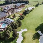 Luftaufnahme eines Resorts mit einem Pool, umliegenden Gärten, kleinen Teichen und einem angrenzenden Golfplatz mit Sandbunkern und grünen Fairways.