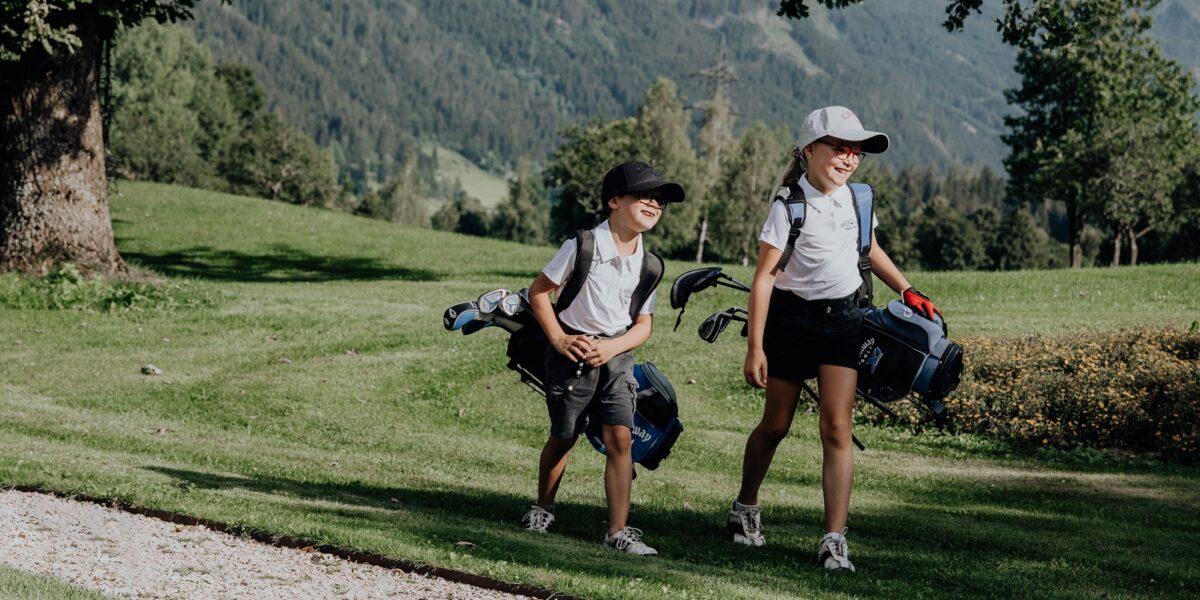 Zwei Kinder mit Mützen und Golftaschen gehen gemeinsam über einen grasbewachsenen Golfplatz mit Bäumen und Bergen im Hintergrund.