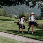 Zwei Kinder mit Mützen und Golftaschen gehen gemeinsam über einen grasbewachsenen Golfplatz mit Bäumen und Bergen im Hintergrund.