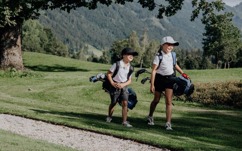 Zwei Kinder mit Mützen und Golftaschen gehen gemeinsam über einen grasbewachsenen Golfplatz mit Bäumen und Bergen im Hintergrund.