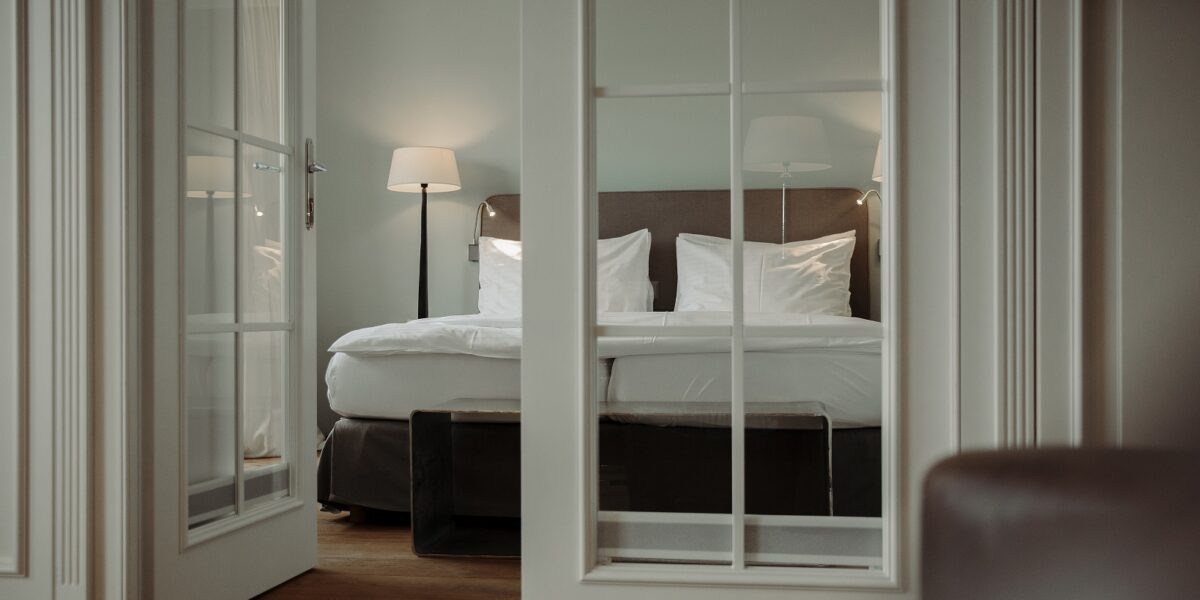 Ein ordentlich gemachtes Bett mit weißer Bettwäsche ist durch offene Glastüren zu sehen, mit einer Stehlampe und sanfter Beleuchtung in einem modernen, minimalistischen Schlafzimmer.