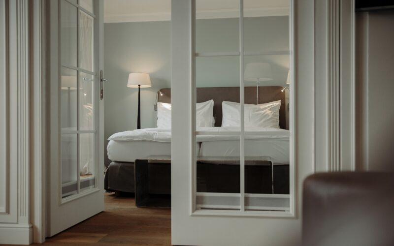 Ein ordentlich gemachtes Bett mit weißer Bettwäsche ist durch offene Glastüren zu sehen, mit einer Stehlampe und sanfter Beleuchtung in einem modernen, minimalistischen Schlafzimmer.