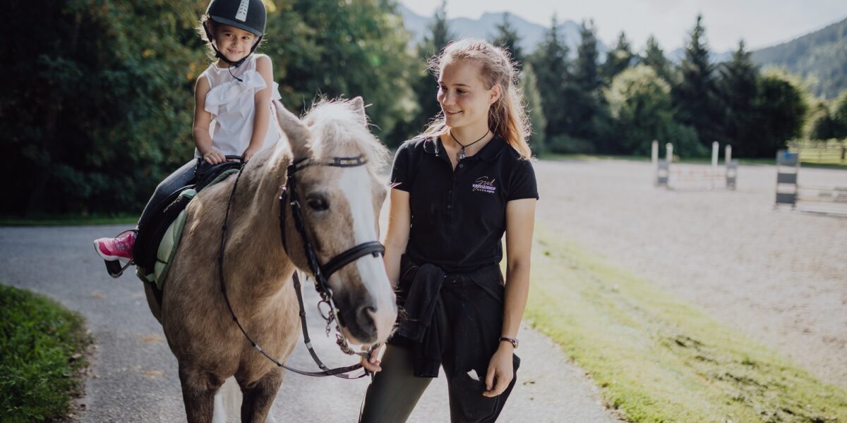 Ein kleines Kind mit einem Helm sitzt auf einem hellbraunen Pferd, während ein älteres Mädchen das Pferd auf einem Reitplatz im Freien führt.