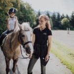 Ein kleines Kind mit einem Helm sitzt auf einem hellbraunen Pferd, während ein älteres Mädchen das Pferd auf einem Reitplatz im Freien führt.