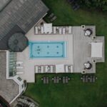 Luftaufnahme eines rechteckigen Außenpools, umgeben von Liegestühlen, Sonnenschirmen und Grünpflanzen, mit einer Person, die im Pool schwimmt.