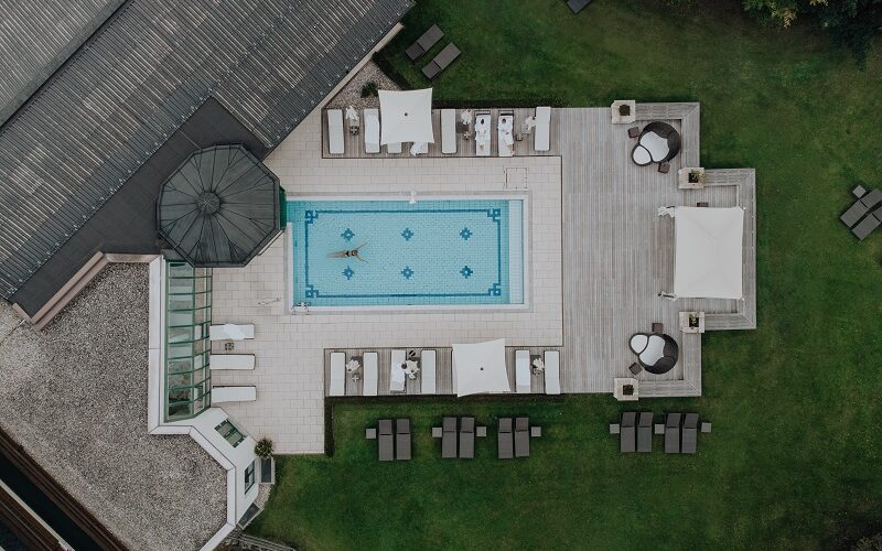 Luftaufnahme eines rechteckigen Außenpools, umgeben von Liegestühlen, Sonnenschirmen und Grünpflanzen, mit einer Person, die im Pool schwimmt.