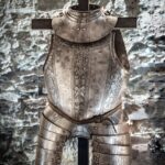 Eine verzierte mittelalterliche Plattenrüstung mit Brustpanzer und Troddeln, ausgestellt auf einem Ständer vor einem Hintergrund aus Steinmauern.