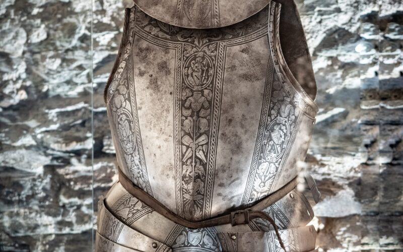 Eine verzierte mittelalterliche Plattenrüstung mit Brustpanzer und Troddeln, ausgestellt auf einem Ständer vor einem Hintergrund aus Steinmauern.