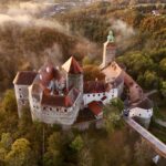 Luftaufnahme einer mittelalterlichen Burg mit roten Dächern und einem hohen Turm, umgeben von dichtem Wald und leichtem Nebel.