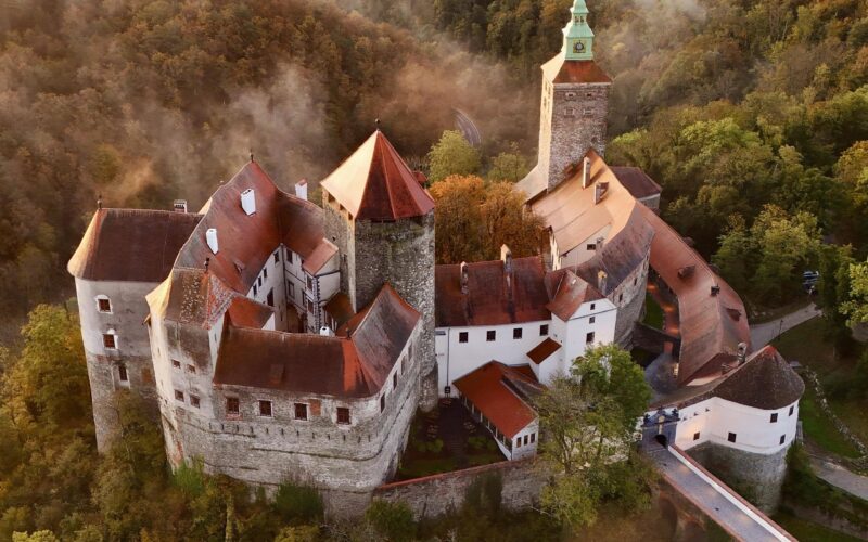 Luftaufnahme einer mittelalterlichen Burg mit roten Dächern und einem hohen Turm, umgeben von dichtem Wald und leichtem Nebel.
