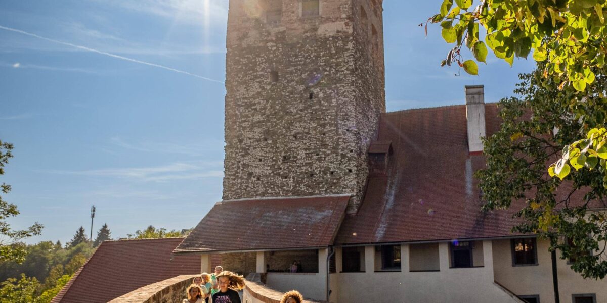 Vier Kinder laufen bei strahlendem Sonnenschein über einen steinernen Weg auf einen historischen Turm zu, mit Bäumen und blauem Himmel im Hintergrund.