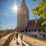 Vier Kinder laufen bei strahlendem Sonnenschein über einen steinernen Weg auf einen historischen Turm zu, mit Bäumen und blauem Himmel im Hintergrund.