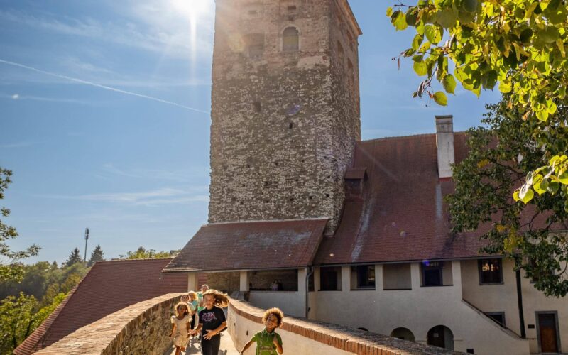 Vier Kinder laufen bei strahlendem Sonnenschein über einen steinernen Weg auf einen historischen Turm zu, mit Bäumen und blauem Himmel im Hintergrund.