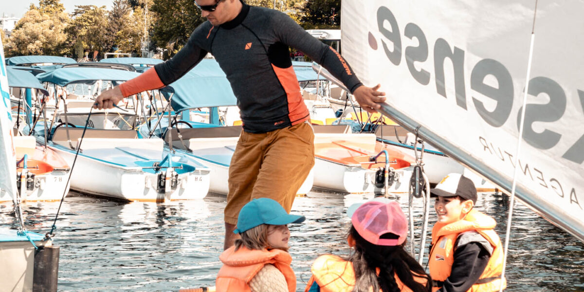 Ein Erwachsener in einem Neoprenanzug steht auf einem Segelboot mit drei Kindern, die Schwimmwesten und Mützen tragen; im Hintergrund sind weitere Boote und Bäume zu sehen.