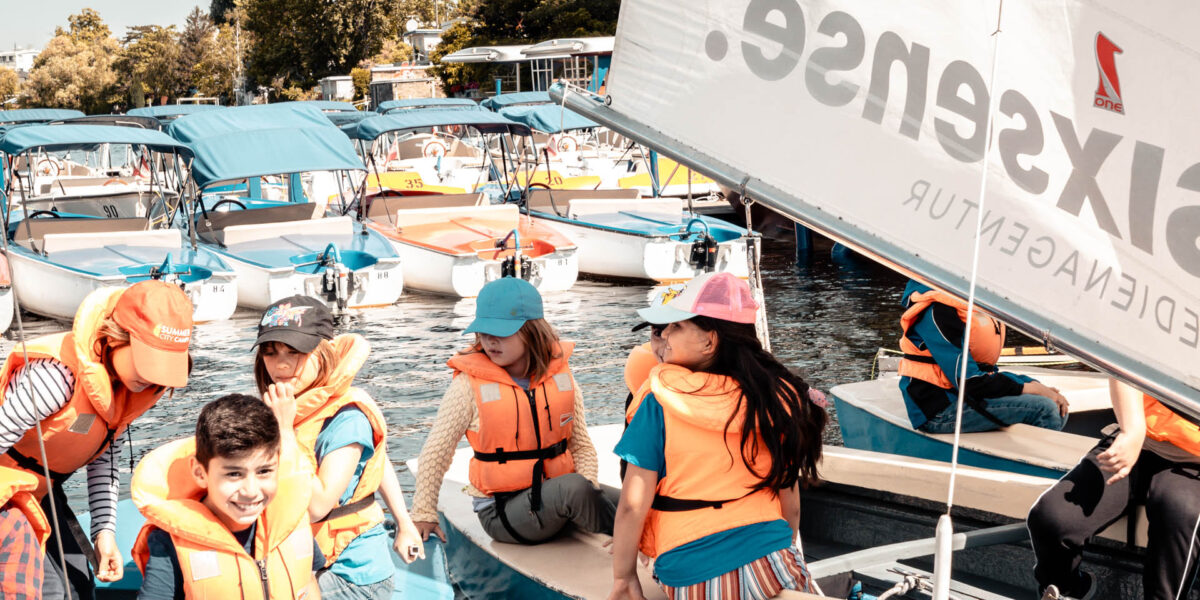 Eine Gruppe von Kindern mit Schwimmwesten sitzt in kleinen Booten auf dem Wasser, im Hintergrund sind andere Boote und Bäume zu sehen.