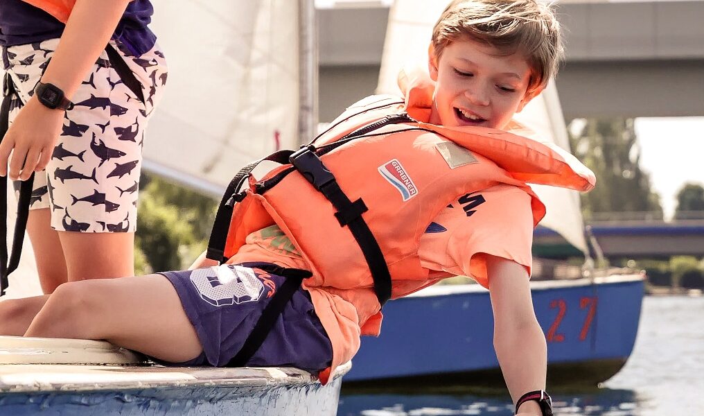 Zwei Kinder mit Schwimmwesten sitzen auf dem Rand eines blauen Segelboots; eines beugt sich vor, um das Wasser zu berühren. Im Hintergrund sind Segelboote und eine Brücke zu sehen.