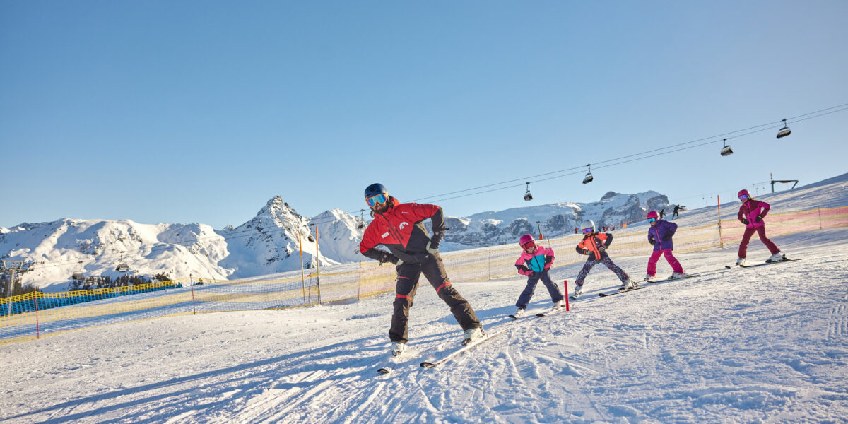 Ein Skilehrer führt eine Gruppe von Kindern in einer Reihe auf einer verschneiten Piste, mit Bergen und Skiliften im Hintergrund.
