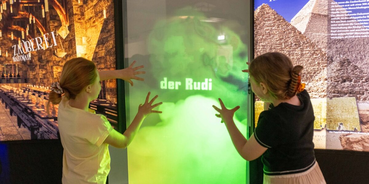 Zwei Kinder interagieren in einer Ausstellung mit einem grün beleuchteten Bildschirm, auf dem die Worte "der Rudi" zu lesen sind, mit Plakaten antiker Stätten im Hintergrund.