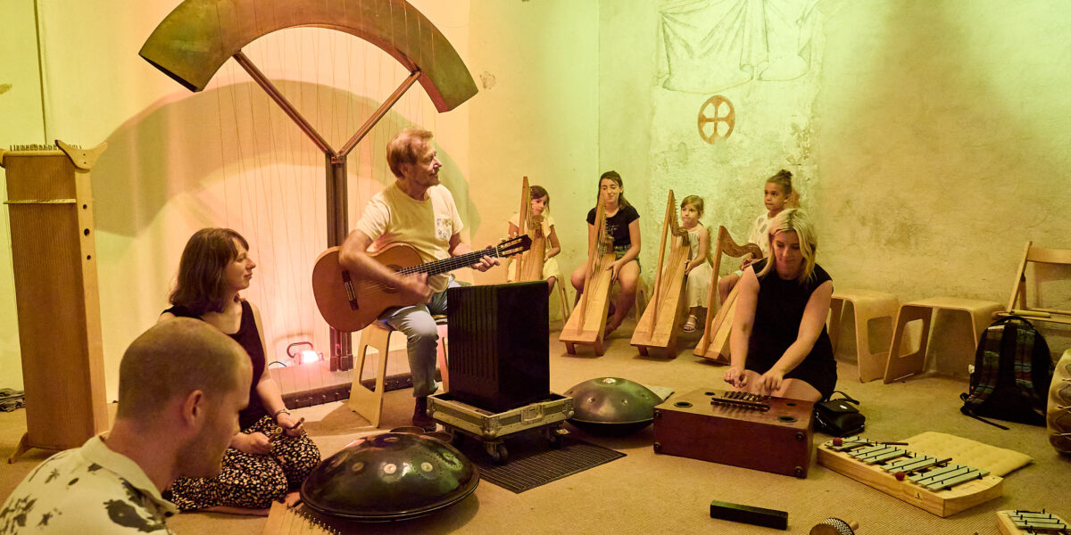 Eine Gruppe von Menschen sitzt in einem Raum und spielt unter warmer gelber Beleuchtung verschiedene Musikinstrumente, darunter eine Gitarre, Xylophone und Handpans.