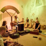 Eine Gruppe von Menschen sitzt in einem Raum und spielt unter warmer gelber Beleuchtung verschiedene Musikinstrumente, darunter eine Gitarre, Xylophone und Handpans.