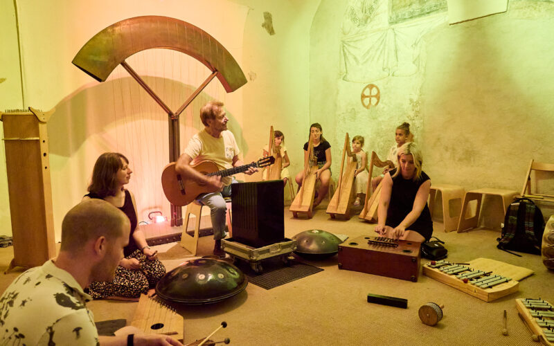 Eine Gruppe von Menschen sitzt in einem Raum und spielt unter warmer gelber Beleuchtung verschiedene Musikinstrumente, darunter eine Gitarre, Xylophone und Handpans.
