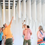 Eine Gruppe von Personen beobachtet und interagiert mit senkrecht hängenden Metallrohren in einem hellen Raum; eine Person schlägt mit einem Hammer auf ein Rohr.