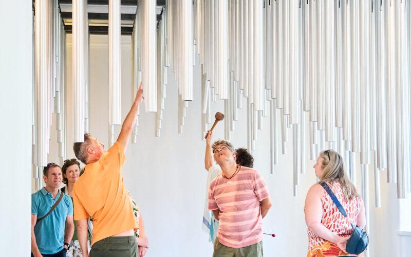 Eine Gruppe von Personen beobachtet und interagiert mit senkrecht hängenden Metallrohren in einem hellen Raum; eine Person schlägt mit einem Hammer auf ein Rohr.