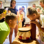 Kinder und Erwachsene versammeln sich um einen Tisch, während ein Mädchen in einem hell erleuchteten Raum an einem hohen Behälter riecht und ein Junge abgedeckte Gläser betrachtet.