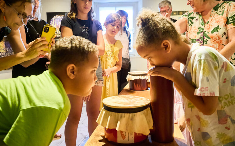 Kinder und Erwachsene versammeln sich um einen Tisch, während ein Mädchen in einem hell erleuchteten Raum an einem hohen Behälter riecht und ein Junge abgedeckte Gläser betrachtet.