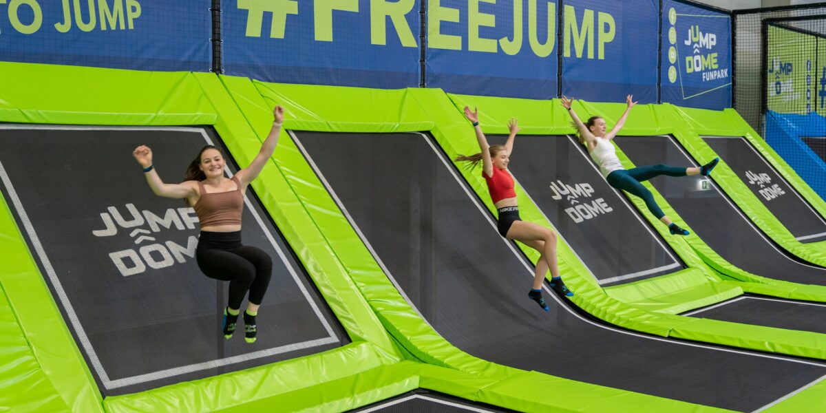 Drei Personen springen auf schrägen Trampolinen in einem Indoor-Trampolinpark mit #FREEJUMP-Schildern im Hintergrund.