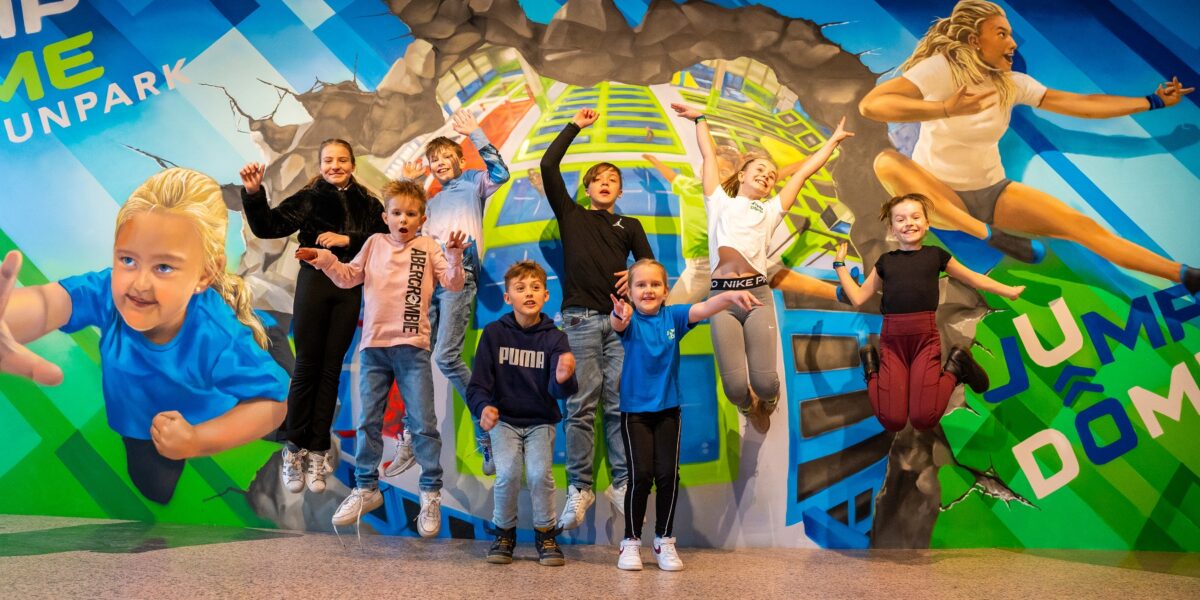 Eine Gruppe von Kindern posiert mitten im Sprung vor einem bunten Wandgemälde in einem Indoor-Sprungpark.