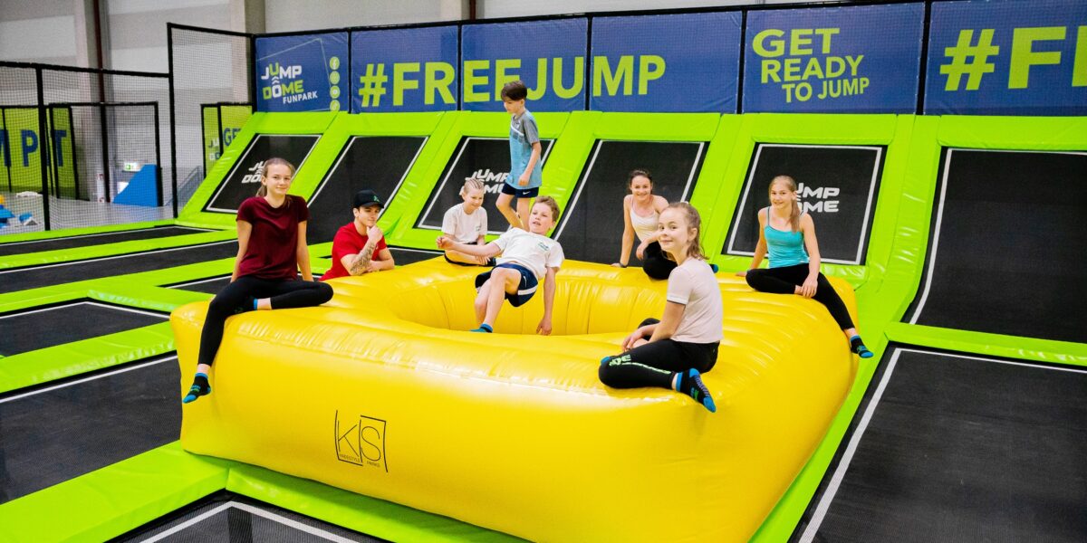 Eine Gruppe von Kindern und Jugendlichen sitzt und steht auf einer großen gelben Hüpfburg in einem Indoor-Trampolinpark, umgeben von Trampolinen und grünen Polstern.