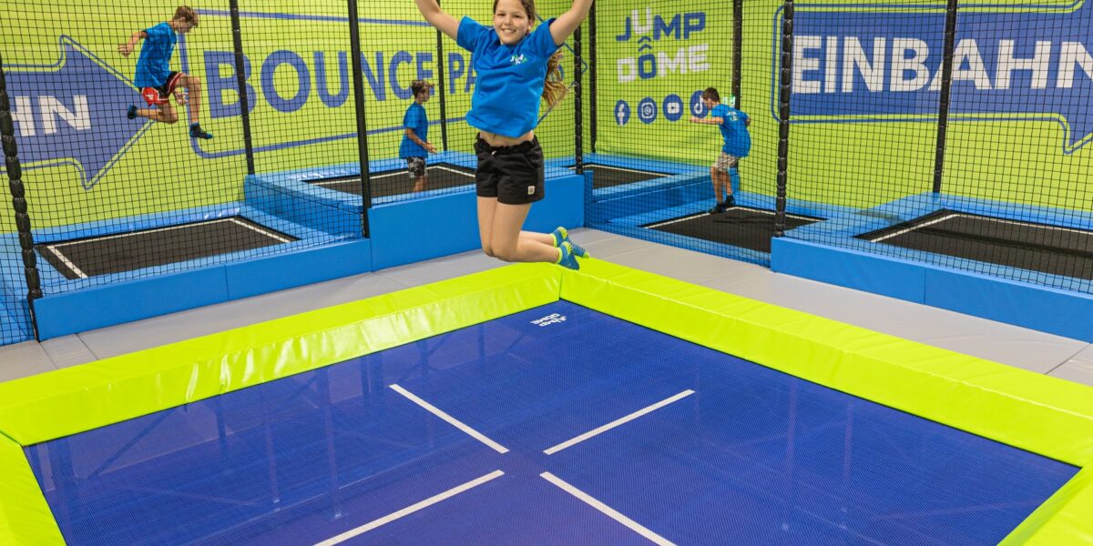 Eine Frau springt mit erhobenen Armen auf einem Trampolin, während Kinder auf anderen Trampolinen in einem Indoor-Trampolinpark spielen.