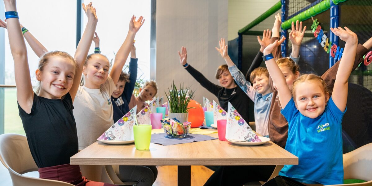 Eine Gruppe lächelnder Kinder sitzt um einen Tisch mit Partyhüten, Tellern und Tassen, hebt die Arme und posiert fröhlich in einer hell erleuchteten Innenumgebung.