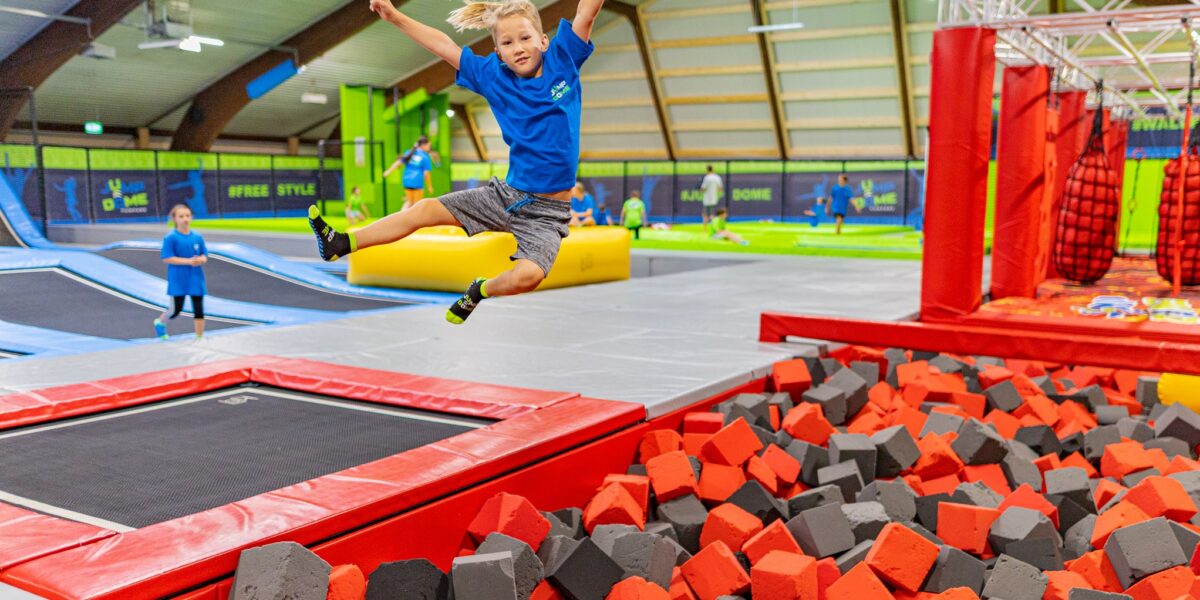 Ein Junge in einem blauen Hemd springt von einem Trampolin in eine Schaumstoffgrube in einem Indoor-Trampolinpark. Im Hintergrund spielen andere Kinder.