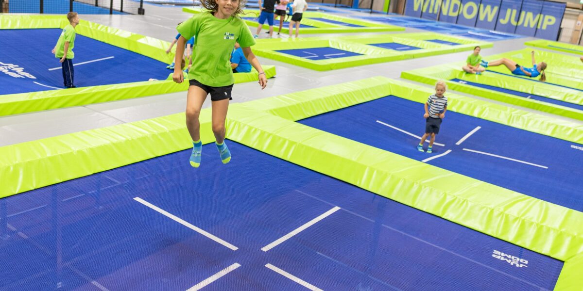Kinder und Erwachsene springen auf blauen und grünen Trampolinen in einem Indoor-Trampolinpark. Ein Mädchen in einem grünen T-Shirt befindet sich in der Luft und lächelt.
