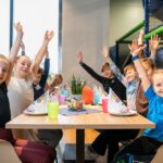 Eine Gruppe Kinder sitzt in einem hell erleuchteten Raum um einen Tisch mit Partydekoration, streckt die Arme in die Höhe und lächelt.