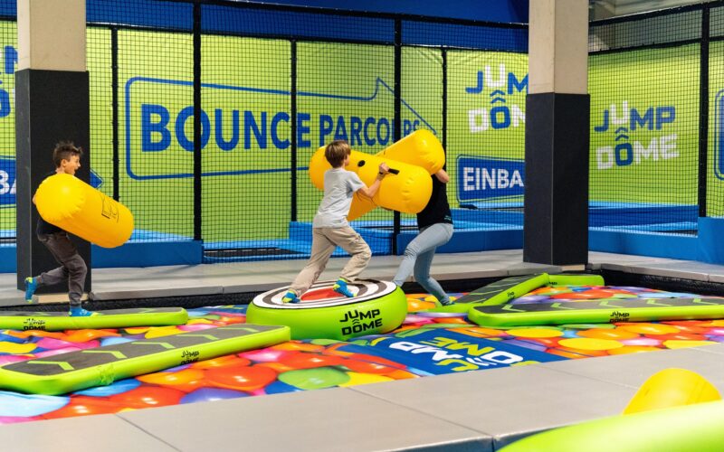Drei Kinder spielen mit großen gelben Polsterstäben auf einer aufblasbaren Plattform im JUMP DOME Salzburg, dem aufregenden Indoor-Trampolinpark.