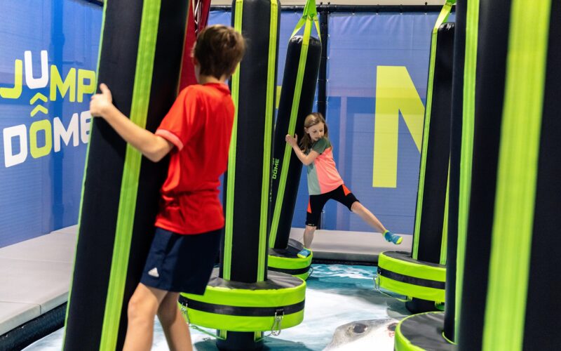Zwei Kinder spielen auf einem weichen Hindernisparcours im Indoor-Aktivitätszentrum JUMP DOME Salzburg und klammern sich an großen gepolsterten Säulen fest, um das Gleichgewicht zu halten.