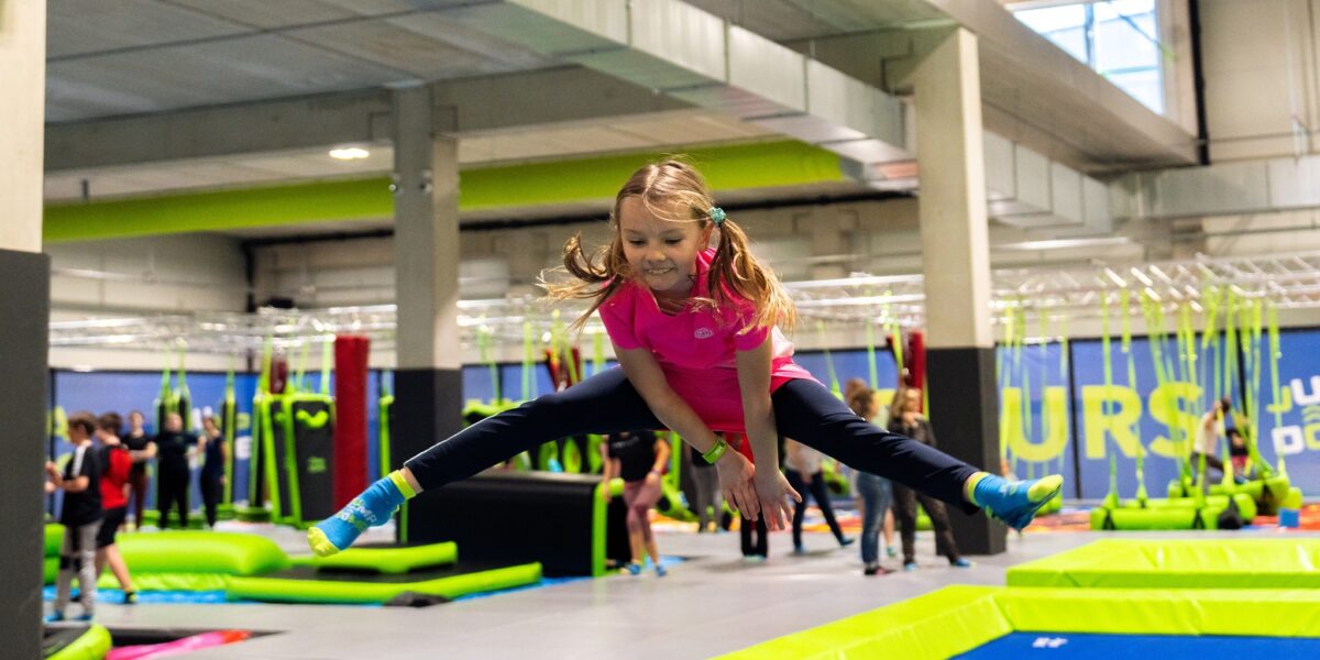 Ein junges Mädchen in Sportkleidung macht einen Spagatsprung über ein Trampolin in einem Indoor-Trampolinpark, im Hintergrund sind andere Leute zu sehen.