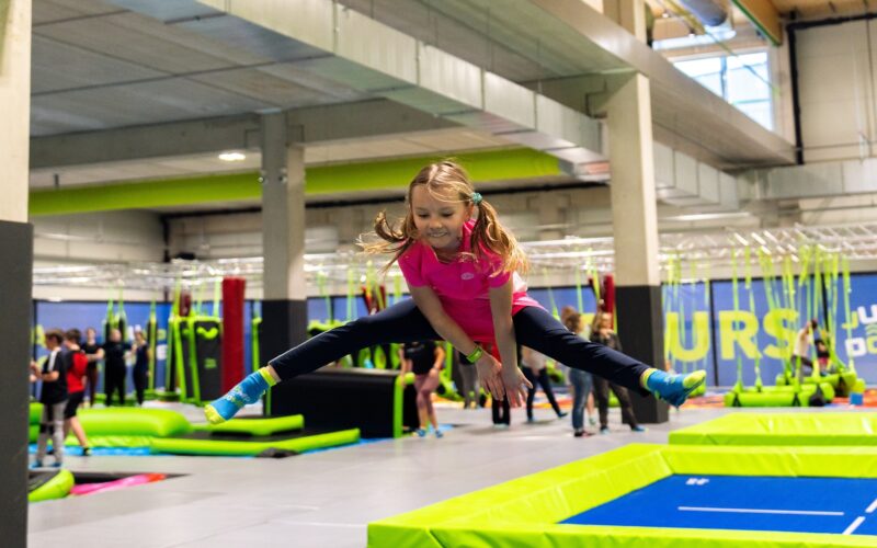 Ein junges Mädchen in Sportkleidung macht einen Spagatsprung über ein Trampolin in einem Indoor-Trampolinpark, im Hintergrund sind andere Leute zu sehen.